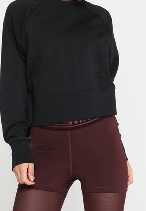 Nærbillede af en person iført en sort langærmet cropped sweatshirt og mørkerøde højtaljede sportsshorts med et logo på taljen og låret.
