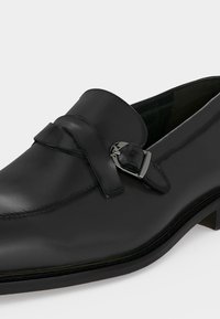 Μαύρο δερμάτινο loafer με ασημένια αγκράφα και χαμηλό στοιβαγμένο τακούνι, εμφανίζεται από πλαϊνή γωνία σε λευκό φόντο.