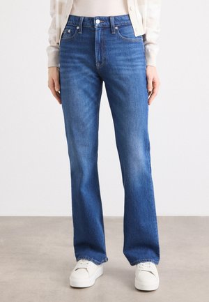 Person trägt mittelblaue Jeans mit geradem Bein, weiße Turnschuhe und eine beige Strickjacke über einem beigen Oberteil und steht auf einem grauen Boden.