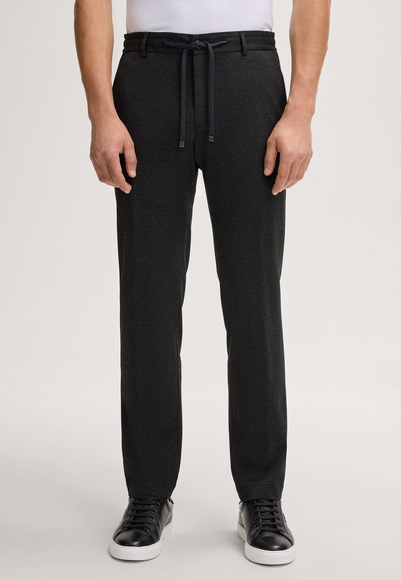 Homme portant un pantalon gris foncé à cordon de serrage et des baskets noires avec des semelles blanches, debout sur un fond uni.