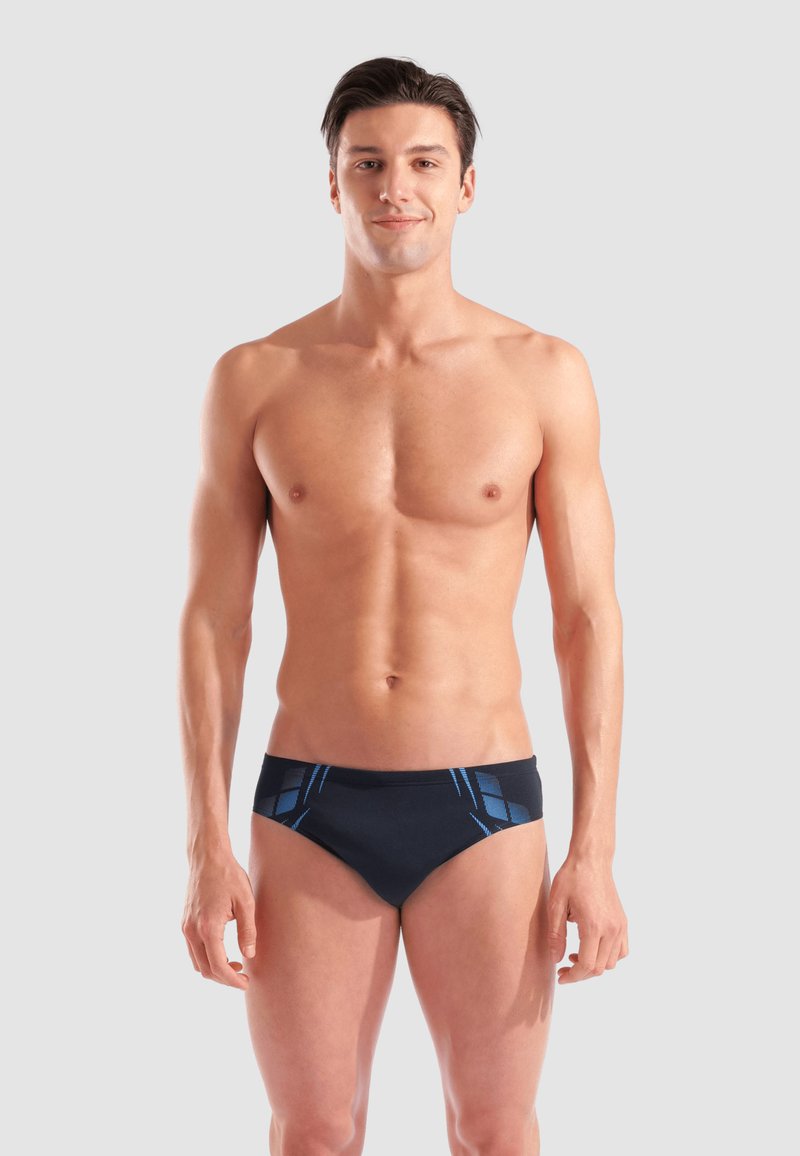 Uomo senza maglietta che indossa slip da bagno neri con accenti geometrici blu, in piedi contro uno sfondo grigio chiaro uniforme, rivolto verso l'obbiettivo.
