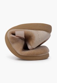 Zapatilla beige con parte superior suave y de felpa, material flexible, forma redondeada y suela de goma beige. Presenta una correa elástica rosa para asegurarla.