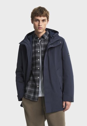 Joven vestido con una chaqueta con capucha azul marino sobre una camisa a cuadros en blanco y negro y pantalones de forro polar beige, de pie contra un fondo simple.