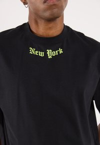 Tricou negru din bumbac cu literă gotică verde care spune "New York", centrat pe piept. Design clasic cu guler rotund.