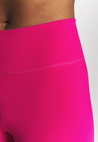 Lähikuvassa ihmisen vyötärö ja lantio, jotka ovat pukeutuneet kirkkaan vaaleanpunaiseen istuviin urheilulegginseihin yksinkertaista vaaleaa taustaa vasten.