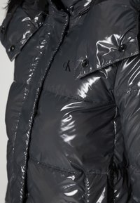 Lśniąca czarna kurtka puchowa z pikowanymi sekcjami, z zatrzaskami, wysokim kołnierzem i małym logo na klatce piersiowej. Charakteryzuje się gładką, błyszczącą teksturą.