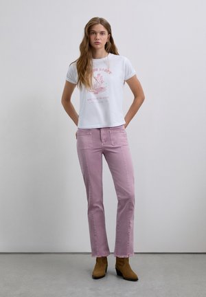 Jeune femme aux cheveux longs, portant un tee-shirt graphique blanc, un jean mauve clair avec des poches avant et des bottes à talons marron.