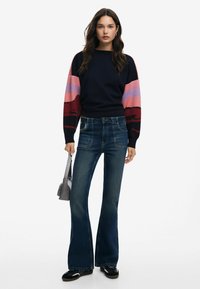 Maglione navy con maniche a strisce rosa e rosse, abbinato a jeans svasati blu scuro. La modella tiene una piccola borsa grigia.