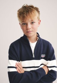 Námornícky pletený sveter s bielymi a čiernymi horizontálnymi pruhmi, so zipsom na krku a nášivkou s logom na hrudi. Mäkká textúra a voľný strih.