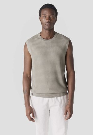 Hombre con un top beige sin mangas de punto texturizado y pantalones blancos, de pie frente a un fondo liso de color gris claro.