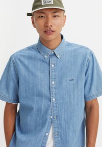 Light blue denim short-sleeve shirt with a button-up collar, featuring a subtle logo, white buttons, and a relaxed fit.