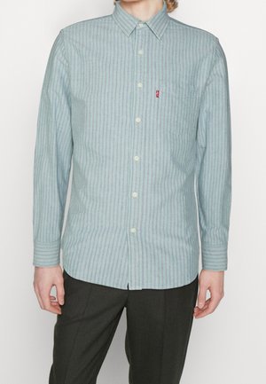 Homme portant une chemise à rayures verticales vert clair et violet avec une poche poitrine et un pantalon noir, debout devant un fond uni.