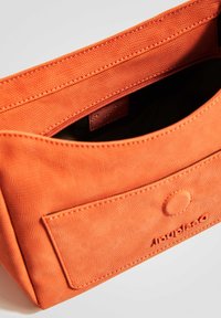 Borsa in tessuto arancione con superficie testurizzata, dotata di tasca frontale, cuciture visibili e logo impresso sul lato. L'interno è nero.