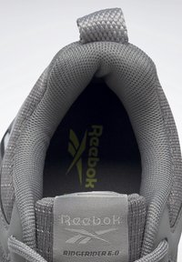 Interiér šedého sportovního boty s texturovanou síťovinou, polstrovaným límcem a logem Reebok na stélce. Přítomné decentní vzory.