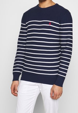 Homme portant un pull bleu marine avec des rayures blanches horizontales et un petit logo rouge sur la poitrine, associé à un pantalon blanc.