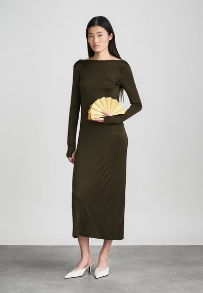 Λαδί στενό φόρεμα με μακριά μανίκια, μέχρι το γόνατο. Το μοντέλο κρατάει ένα κίτρινο φανελάκι, σε συνδυασμό με λευκά ψηλοτάκουνα παπούτσια με δαντέλα.