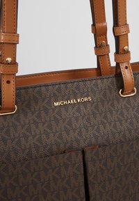 Sac fourre-tout marron en tissu texturé, avec des accessoires en métal doré, des anses en cuir et un logo "MICHAEL KORS" bien en vue.