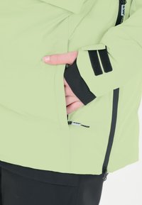 Vihreä vedenpitävä takki, jossa on vetoketjullinen tasku, mustat yksityiskohdat, tarranauha-aksentit ja sileä pinta. Sopii ulkoilutoimintoihin.