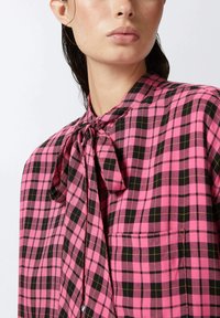 Blusa a quadri rosa e nera con colletto a fiocco, dotata di tasca frontale e realizzata in un tessuto morbido e testurizzato.
