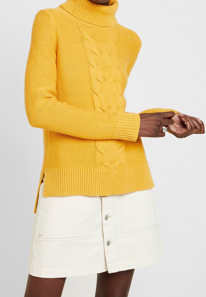 Gelber, grob gestrickter Rollkragenpullover mit strukturierter Stoffoberfläche, gerippten Bündchen und seitlichen Schlitzen. Kombiniert mit einem weißen Jeansrock mit Knöpfen.