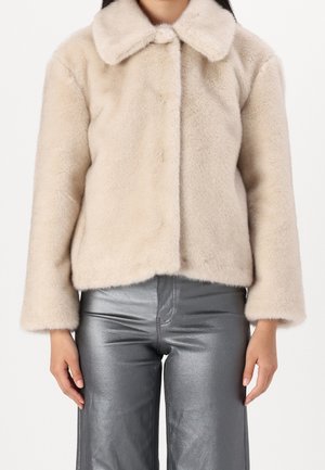 Femme portant une veste en fausse fourrure beige avec un large col et un pantalon argenté brillant taille haute, debout devant un fond uni.