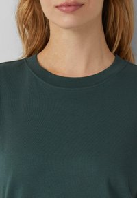 T-shirt vert foncé avec un col rond et un tissu lisse et doux. Comprend des coutures discrètes le long du col et une coupe décontractée.