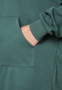 Zelená bavlněná mikina s přední kapsou, žebrovanými manžetami a měkkým, texturovaným povrchem. Jednoduchý design s minimálními ozdobami.