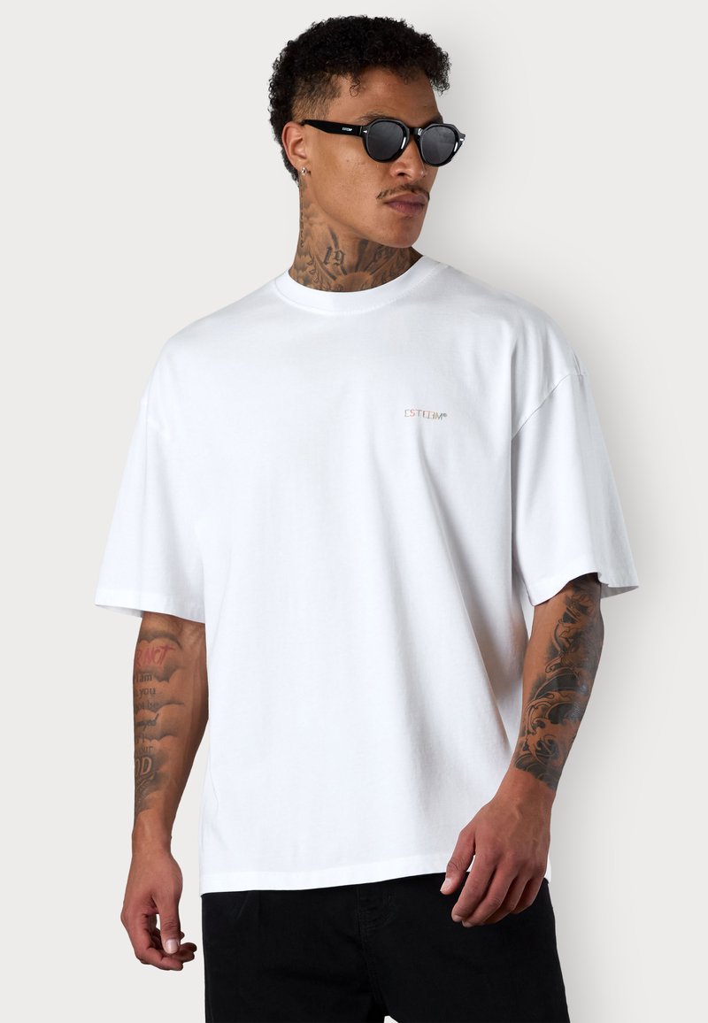 White cotton t-shirt with a relaxed fit, round neckline, and short sleeves. Features small "ESTEEM" logo in pink on the chest. Black pants and sunglasses.