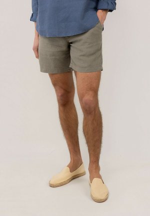Hombre vestido con pantalones cortos beige, camisa azul de manga larga y alpargatas crema sin cordones, de pie con una mano en el bolsillo.