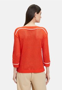 Roter Strickpullover mit einem lockeren, offenen Strickdesign, Three-Quarter-Ärmeln und weißen Akzentstreifen am Ausschnitt und den Bündchen.