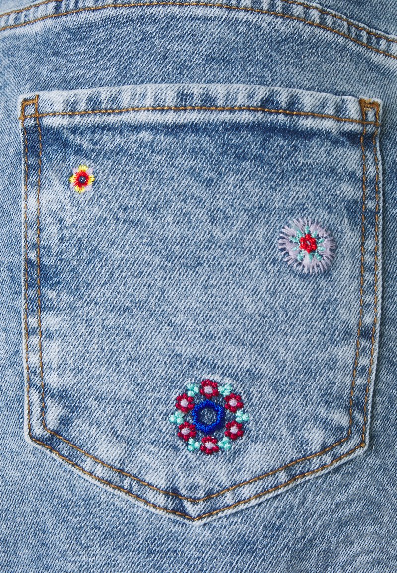 Denimtasche mit mehrfarbiger Blumenstickerei: drei verschiedene Muster in Rot, Blau und Weiß auf hellblauem Stoff mit sichtbaren Nähten.