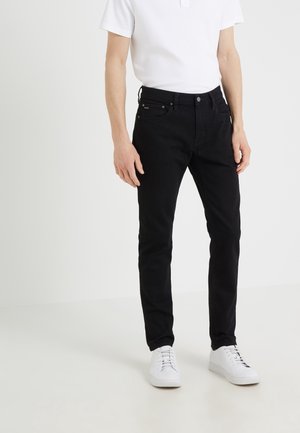 Zwarte jeans met een slim fit, denim stof, vijf zakken en subtiele stikdetails, gecombineerd met witte sneakers en een wit poloshirt.