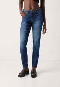 Jeans de mezclilla azul con corte recto, dos bolsillos frontales y cierre de dos botones. Llevados con sandalias negras de tacón cuadrado.