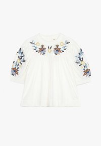 Blouse en coton blanc avec des motifs floraux brodés en orange, bleu et jaune sur les manches, dotée d'un col rond et d'une coupe évasée.