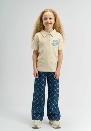 Polo shirt beis con bordado 'Ciao Bella', combinado con pantalones de mezclilla oscuros de pierna ancha con patrones florales y un ajuste relajado. Zapatillas ligeras.