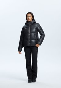 Chaqueta de plumas negra con capucha, con cierre de cremallera y detalles de costura. Combinada con pantalones negros y calzado oscuro.