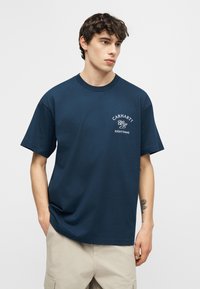 Νέος άνδρας που φοράει μπλουζάκι Carhartt WIP σε ναυτικό μπλε και μπεζ σορτς, στέκεται με τα χέρια στις τσέπες, μπροστά σε απλό ανοιχτό φόντο.