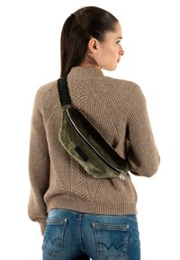 Banane olive en matériau doux et texturé avec une fermeture éclair argentée et une sangle noire réglable avec un logo. Portée dans le dos.