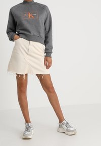 Felpa grigia con collo rotondo e logo Calvin Klein, abbinata a una gonna in denim bianca con orlo sfilacciato e sneakers grigie con dettagli.