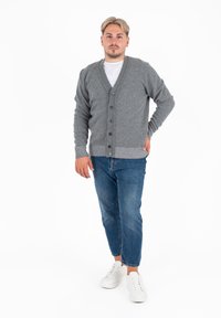Cardigan grigio a maglia con scollo a V, chiusura a bottoni e polsini a coste, indossato sopra una maglietta bianca, abbinato a jeans blu e sneaker bianche.