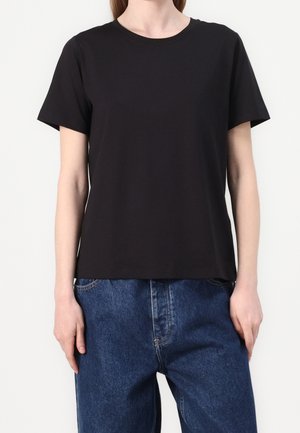 Person trägt ein schlichtes schwarzes T-Shirt mit kurzen Ärmeln, das über locker sitzenden blauen Jeans aus Denim getragen wird, und steht vor einem weißen Hintergrund.