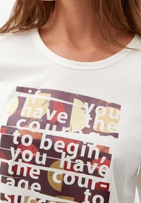 Weißes Baumwoll-T-Shirt mit einem grafischen Design, das Text in verschiedenen Farben zeigt: burgunderrot, gelb und orange, sowie ein geschichtetes gestreiftes Muster.