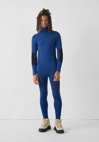 Top atletico a maniche lunghe blu con accenti neri e dettagli testurizzati, abbinato a leggings coordinati e stivali da trekking color beige chiaro.