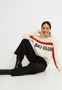 Suéter crema con "SKI CLUB" en negro, franja roja atravesando el pecho, tela texturizada. Combinado con pantalones negros de pierna ancha y botas negras.