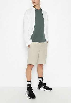 Camicia bianca a maniche lunghe, t-shirt verde, pantaloni corti beige, sneaker nere con dettagli bianchi e calze a strisce nere con logo "AIX".