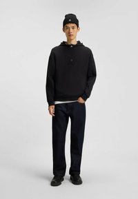 Felpa nera con logo in rilievo, abbinata a jeans blu scuro. Gli accessori includono un berretto di maglia nero e scarpe nere. Design semplice e moderno.