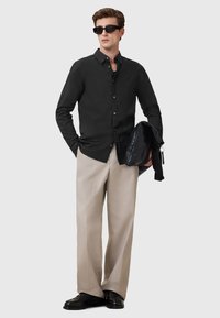 Mann mit schwarzem Hemd, beigen weiten Hosen, schwarzen Schuhen und Sonnenbrille, der eine schwarze Lederhandtasche vor einem schlichten Hintergrund hält.