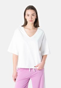 Balta V formos iškirpte t-shirt su trumpomis rankovėmis, pagaminta iš lengvo audinio. Dėvima kartu su rožinėmis kelnėmis, kurios turi baltas šonines juostas kontrastui.