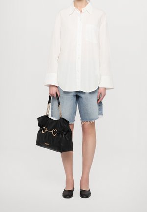 Persoon die een wit shirt met lange mouwen draagt, lichtblauwe denim shorts tot op de knieën, zwarte platte schoenen en een zwarte handtas met gouden hartvormige accenten vasthoudt.