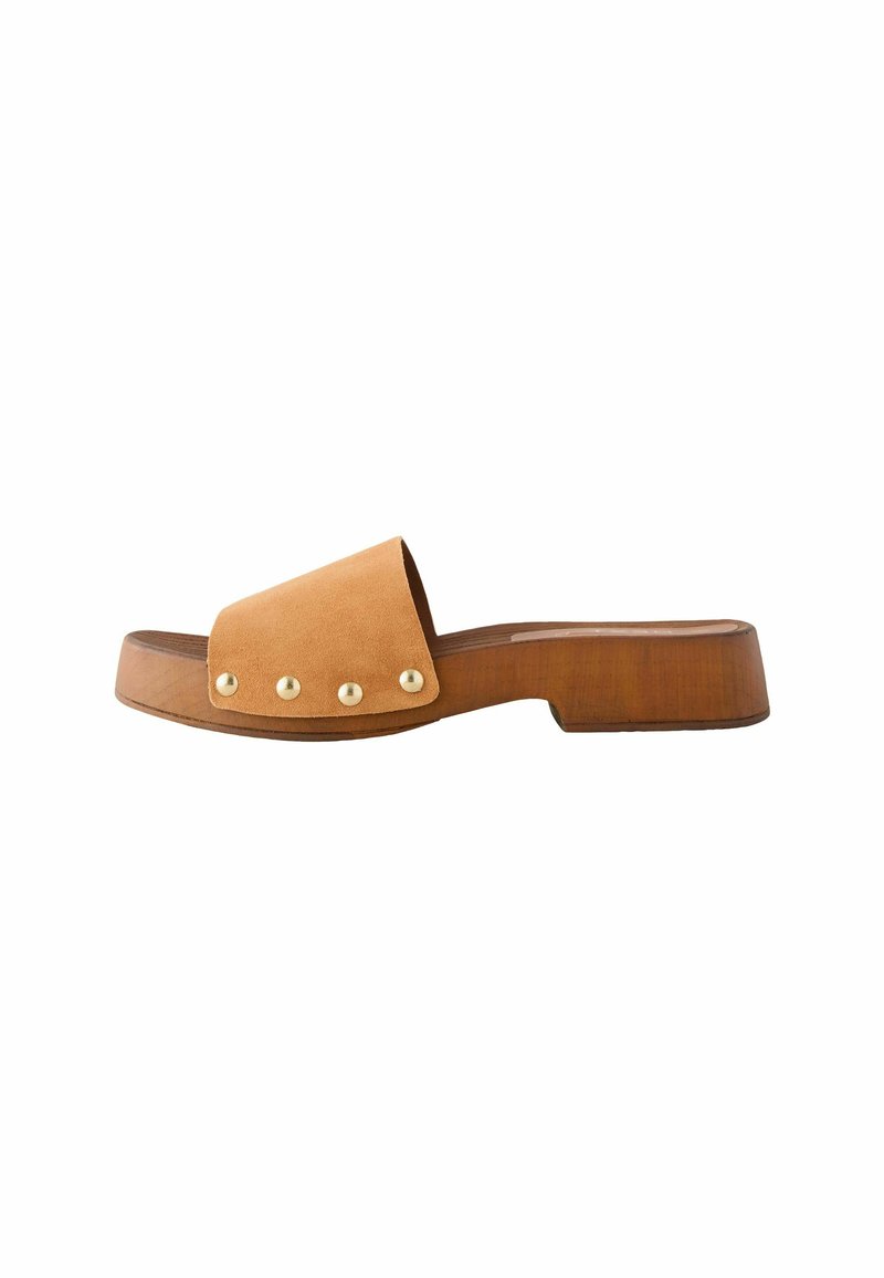Sandale glissière en daim couleur tan avec une large bandoulière et trois clous dorés. Elle dispose d'une semelle en bois épaisse avec un léger talon et un bout arrondi.